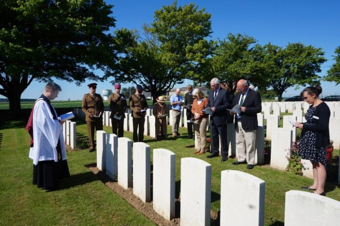 A group of people, including military personnel, attend a solemn ceremony at a cemetery, where rows of white headstones honor the Great War soldiers under a clear blue sky.