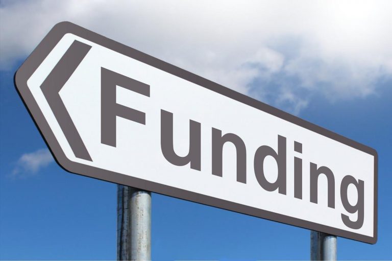 A road sign with the words "New Funding" and an arrow pointing to the left stands against a cloudy sky, hinting at fresh opportunities for military support.