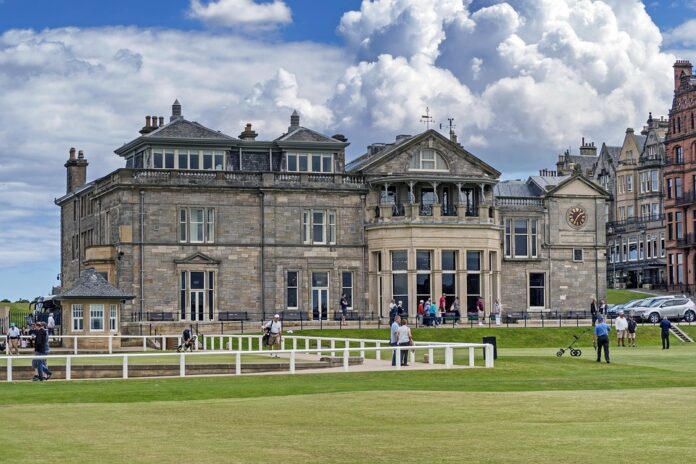 The historic stone clubhouse at the golf course, featuring a clock tower, is surrounded by green lawns and people. As golfers gather, the atmosphere feels like a scene from 