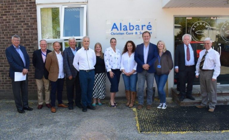 A group of thirteen people standing in front of a building with a sign that reads "Alabaré Christian Care & Support," dedicated to tackling homelessness with innovative self-build projects.
