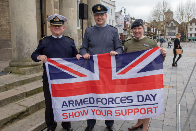 Three people in military uniform hold a Union Jack flag with "Armed Forces Day Show Your Support" printed on it, standing on a street lined with buildings and trees, celebrating the event with pride.