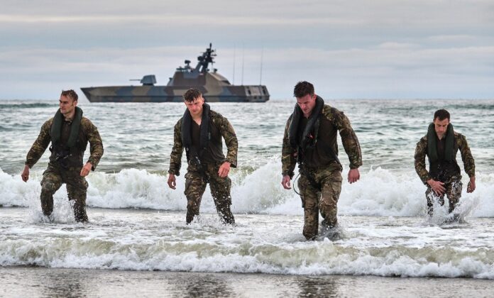Four soldiers in camouflage uniforms walk through the surf on a beach, marking the complete first phase of their Baltic deployment, with a military ship visible in the background.