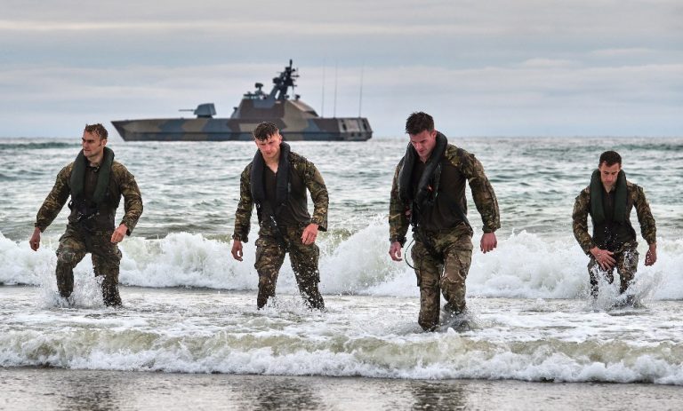 Four soldiers in camouflage uniforms walk through the surf on a beach, marking the complete first phase of their Baltic deployment, with a military ship visible in the background.