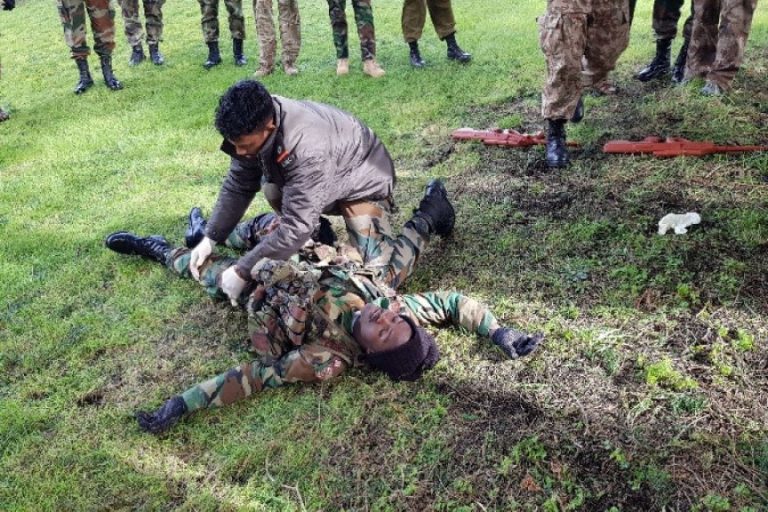 During a UN training session, a person in civilian clothing demonstrates first aid on a soldier lying on grass, emphasizing the importance of collaboration. Others stand nearby observing, unified in their efforts to learn and support one another.