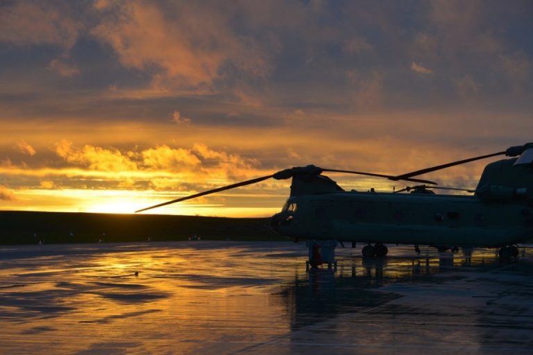 A military helicopter is parked on a wet runway in Croatia, silhouetted against a dramatic sunset with orange and yellow hues, preparing for airborne exercises as troops prepare for the mission ahead.