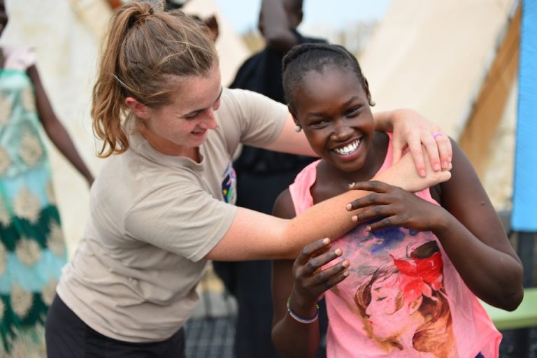 Two women engage in playful interaction, one wearing a beige shirt and the other in a pink top with a printed design. Amidst the blurred background of people, their camaraderie subtly highlights the power of connection and prevention in conflict situations.