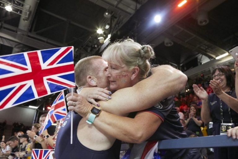 In a crowded indoor venue, partners warmly embrace, one clutching a Union Jack flag—a moment of unity that resonates with the spirit of the Invictus Trials.