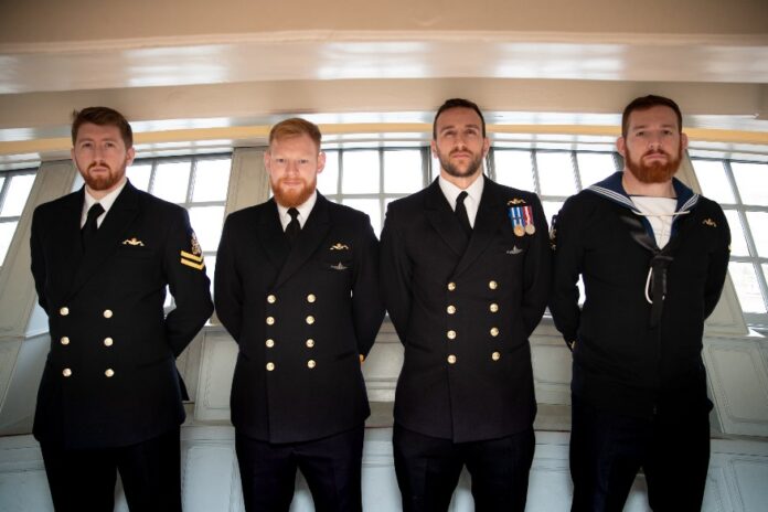 Four men in navy uniforms stand in a line against a windowed wall, looking straight ahead, during the Oardacious Boat unveiling ceremony.
