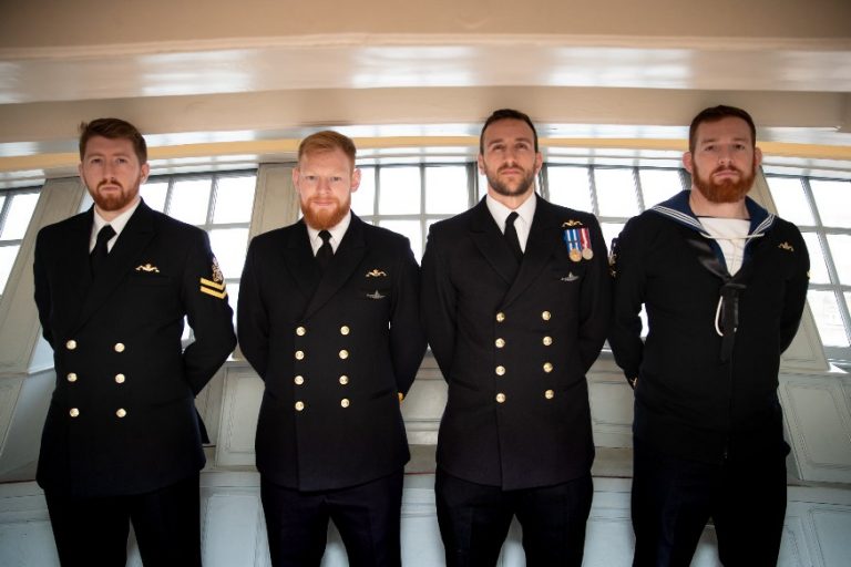 Four men in navy uniforms stand in a line against a windowed wall, looking straight ahead, during the Oardacious Boat unveiling ceremony.