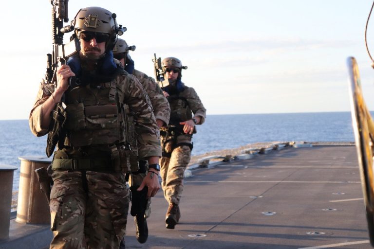 Three military personnel in camouflage and gear walk on the deck of an RN warship, focused on preventing human trafficking, with the ocean stretching endlessly in the background.