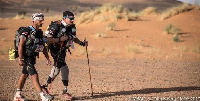 Two athletes traverse the desert terrain, showcasing resilience as they run in their marathon gear. Among them, a determined double amputee uses trekking poles, pushing boundaries and inspiring all as they race through the challenging landscape.