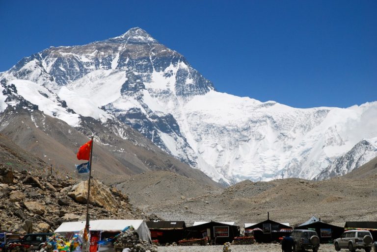 Snow-covered Mount Everest under a clear blue sky offers breathtaking sights, with tents and vehicles dotting the foreground, as if an Army man were leading a peaceful expedition amidst nature's grandeur.