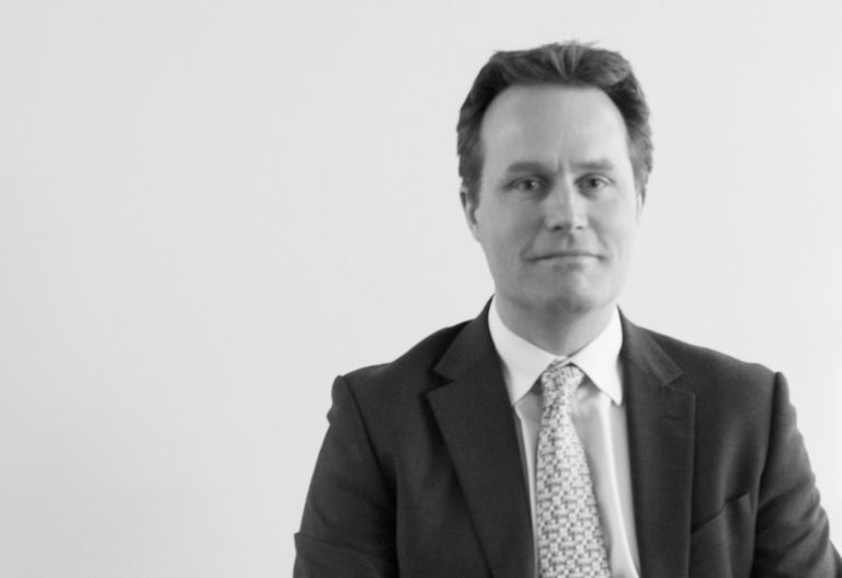 Black and white photo of a man in a suit and tie, sitting against a plain background, exuding the calm confidence of an Ops Director adept at managing combat stress.