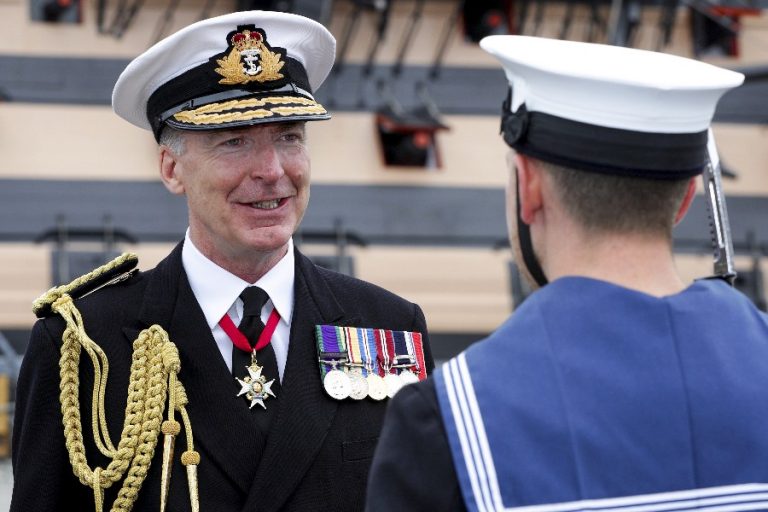 Two naval officers in uniform face each other. The officer on the left, possibly the newly appointed First Sea Lord, wears a decorated uniform with medals and a peaked cap.