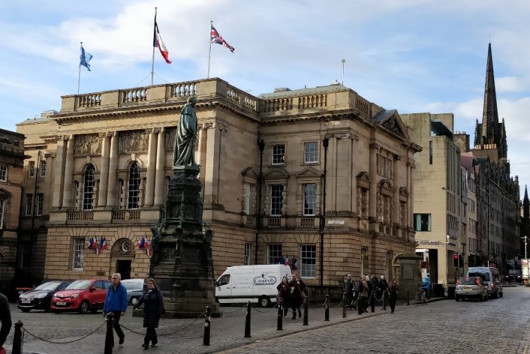 The historic building, adorned with flags representing the Auld Alliance, stands proudly with a statue in front. Pedestrians stroll along the cobblestone street, while a church spire rises in the background on this clear day.