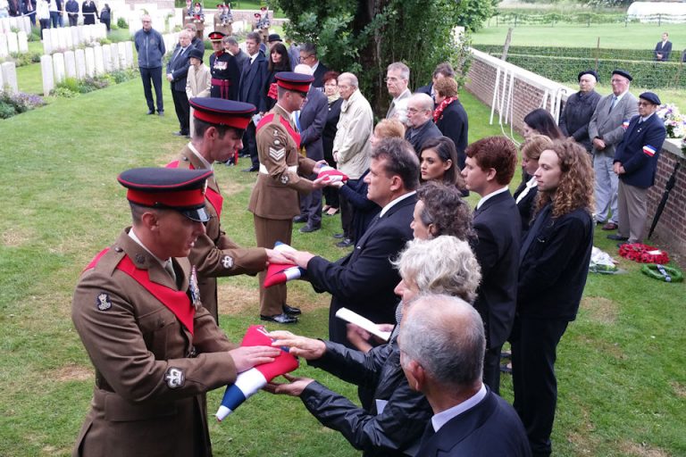Families receive folded flags from military personnel in uniform during a solemn outdoor funeral ceremony, with attentive onlookers present.