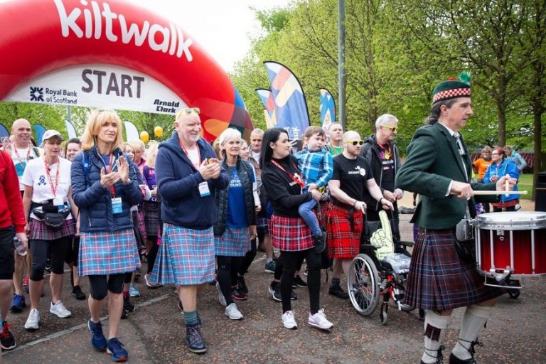 Kiltwalkers, including a drummer and participants in wheelchairs, proudly start the walk under a "kiltwalk" banner in the park. United for a cause, they aim to raise £12k together.
