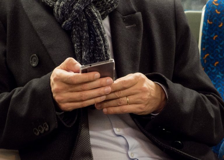 A person in a coat and scarf sits on public transport, holding a smartphone with both hands, likely checking the latest updates on the Veterans' Lottery.