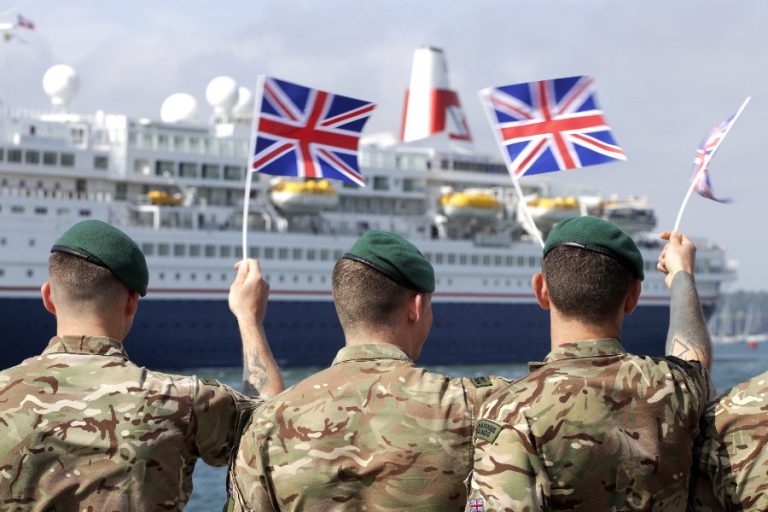 Three marines in camouflage hold British flags proudly in front of a cruise ship, paying homage to their forefathers who bravely landed on D-Day.
