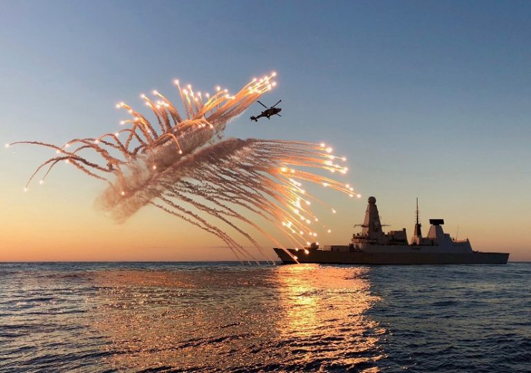 During a stunning sunset, a naval ship glides across the sea as a helicopter releases flares, capturing the magic and majesty of the moment. This breathtaking scene is reminiscent of entries in the prestigious RN Photographic Competition.