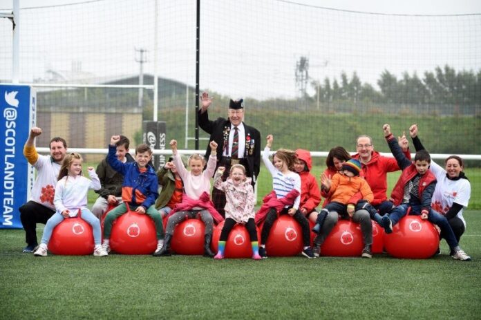 A group of children and adults sitting on red space hoppers in a row on a sports field, smiling and raising their arms in celebration, ready to get bouncing with joy.