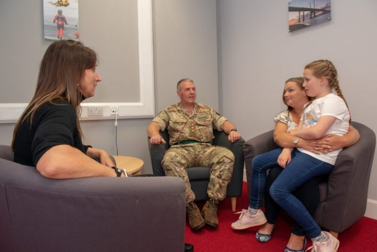 A man in military uniform sits with three people: a woman, another woman holding a child, and a third woman seated across the room. They appear to be engaged in conversation, possibly discussing the community's tribute to local reservists.