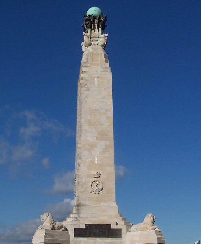 Against a backdrop of blue sky, this tall stone monument commemorates veterans with its sphere and statues at the top, accompanied by two lion figures at the base, evoking the valor of D-Day heroes.