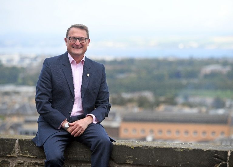 A man in a suit and pink shirt, who is rumored to be the city's Veterans Commissioner, sits on a stone wall with a blurred cityscape in the background.