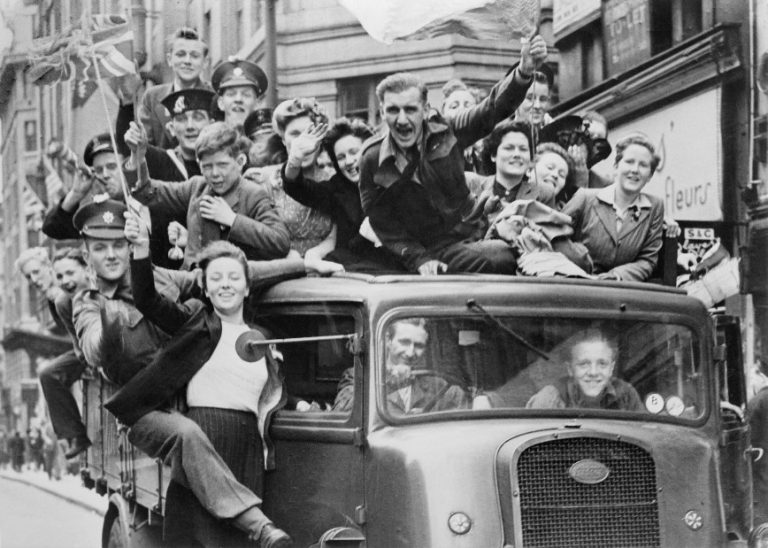A group of people, including uniformed individuals, cheerfully ride on top of and inside a vehicle on a city street during the Bank Holiday, waving flags.