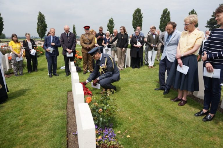 A group of people stands around a cemetery, paying homage as a person in uniform kneels by a gravestone adorned with flowers. The scene is solemn and respectful, remembering the Welsh Brothers who have been laid to rest.