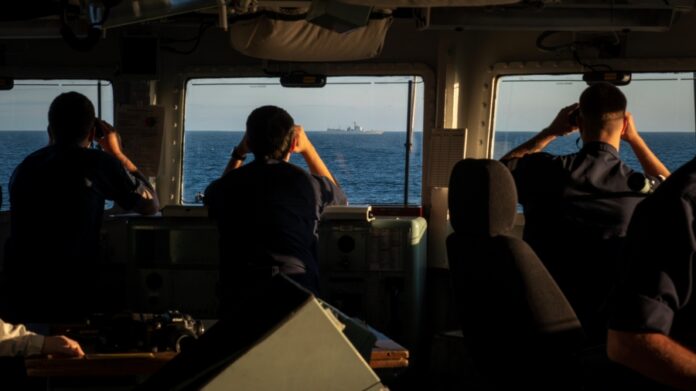 Silhouetted figures on a frigate's bridge use binoculars to monitor a Chinese ship on the horizon at sea during daylight.