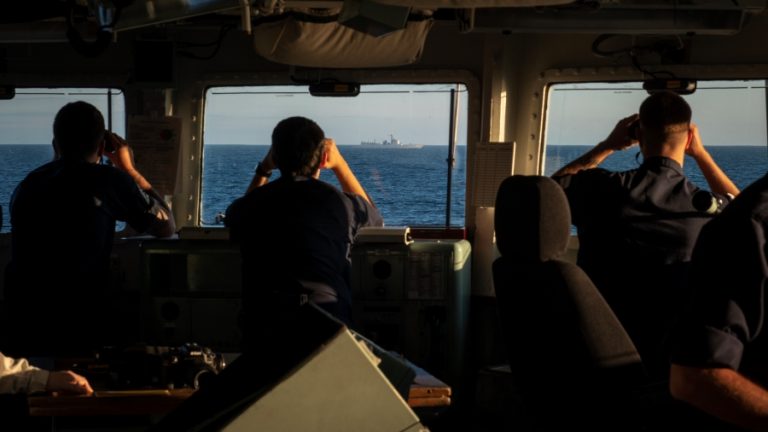 Silhouetted figures on a frigate's bridge use binoculars to monitor a Chinese ship on the horizon at sea during daylight.