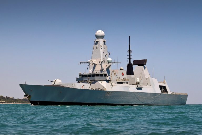 A naval destroyer, participating in a major exercise, sails on calm blue waters under a clear sky, with its radar tower and various equipment prominently visible on deck.