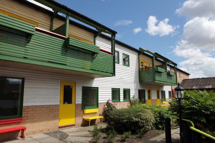 A two-story building with green and yellow accents, featuring balconies and windows, stands proudly. In front, a garden with shrubs and a lamp post embraces the scene under a partly cloudy sky, symbolizing hope for veterans seeking secure employment.
