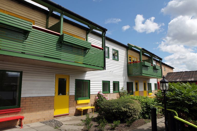 A two-story building with green and yellow accents, featuring balconies and windows, stands proudly. In front, a garden with shrubs and a lamp post embraces the scene under a partly cloudy sky, symbolizing hope for veterans seeking secure employment.