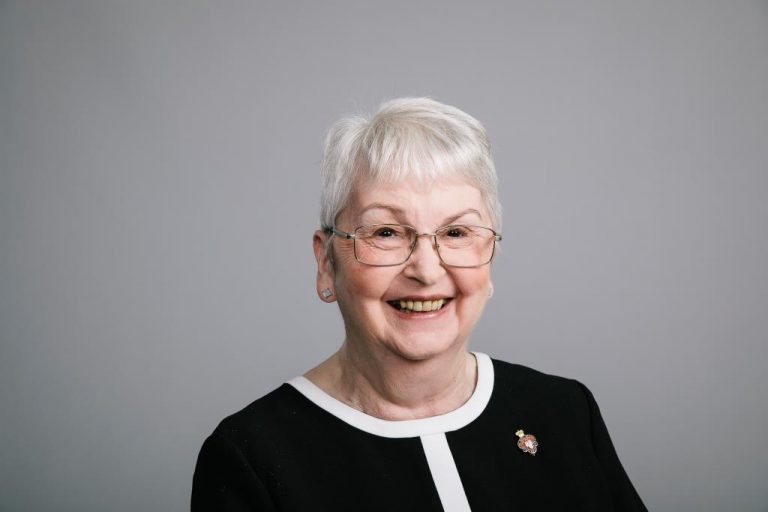Elderly woman with short white hair and glasses, smiling, wearing a black top with a white collar against a plain gray background. As the first female national chairman of the Legion elects, her poised demeanor reflects her groundbreaking achievement.