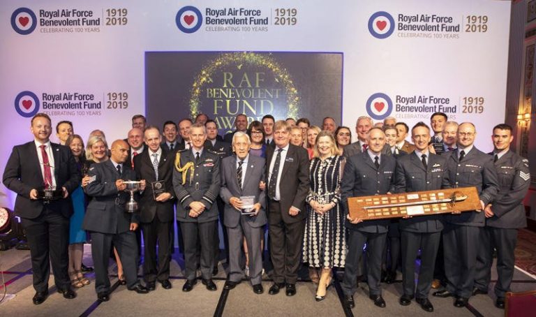 A group of supporters in formal attire pose together on a stage at the celebrated event honoring the Royal Air Force Benevolent Fund's 100th anniversary.