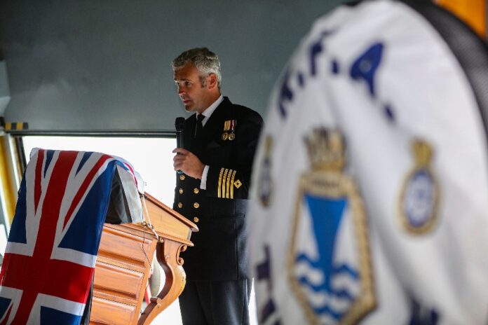 At the Armed Forces Expo in Plymouth, a man in a Royal Fleet Auxiliary naval uniform speaks at a podium draped with a Union Jack flag, while a blurred flag featuring a crest flutters in the foreground.