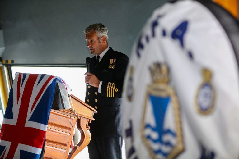 At the Armed Forces Expo in Plymouth, a man in a Royal Fleet Auxiliary naval uniform speaks at a podium draped with a Union Jack flag, while a blurred flag featuring a crest flutters in the foreground.