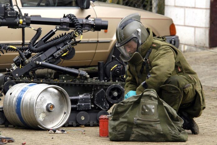 A bomb disposal technician, one of our former Bomb Disposal Heroes, kneels in a protective suit beside essential equipment and a robotic device near a barrel outdoors.