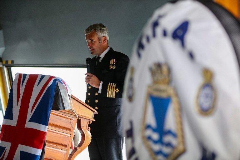 A uniformed officer speaks at a podium draped with a British flag, like the final piece of a jigsaw puzzle completing the picture. Another uniformed individual stands in the foreground, attentive and poised.