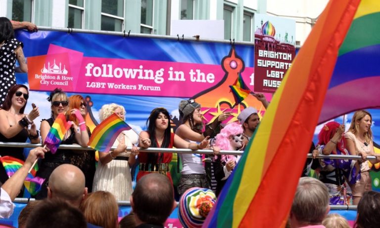 People on a parade float waving rainbow flags at a pride event celebrate inclusivity. A banner reads "following in the Brighton & Hove City Council LGBT Workers Forum," highlighting the council's commitment to progressive LGBT policy and community support.