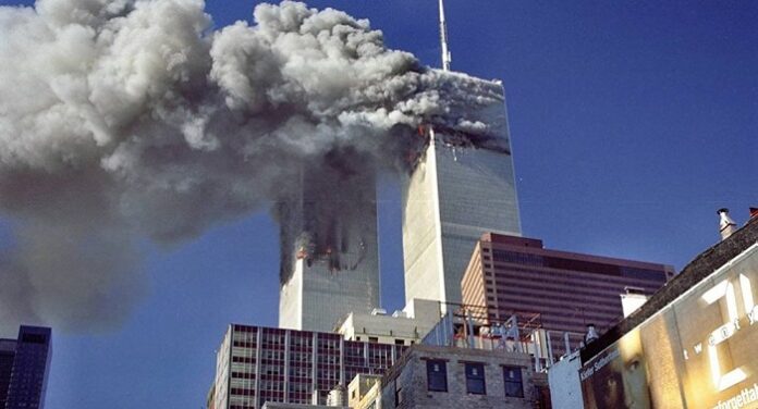 Two skyscrapers emitting large plumes of smoke from the upper floors against a clear blue sky, with other buildings visible in the foreground, serve as haunting reminders of the day the world changed forever.
