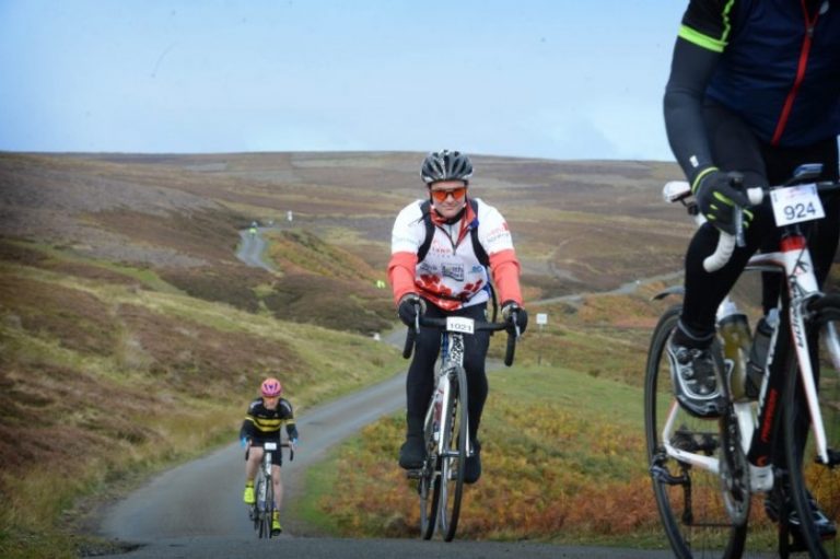 Cyclists ride uphill on a rural road during a sportive success event, sporting numbered jerseys and safety gear, surrounded by rolling hills.