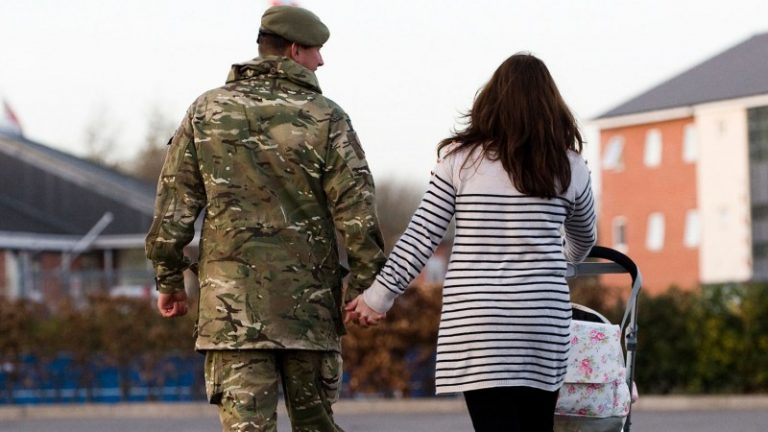 A person in a military uniform and another in a striped sweater walk hand in hand, proudly showcasing the unity of Armed Forces families as they push a floral-patterned stroller along the paved path.
