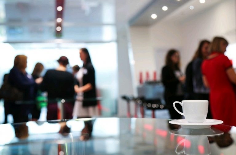 A coffee cup sits on a glass table in the foreground, as a group of people engages in conversation against a modern, blurred backdrop. Their chat revolves around the flexible approach to improving employability, making opportunities out of happenstance encounters.