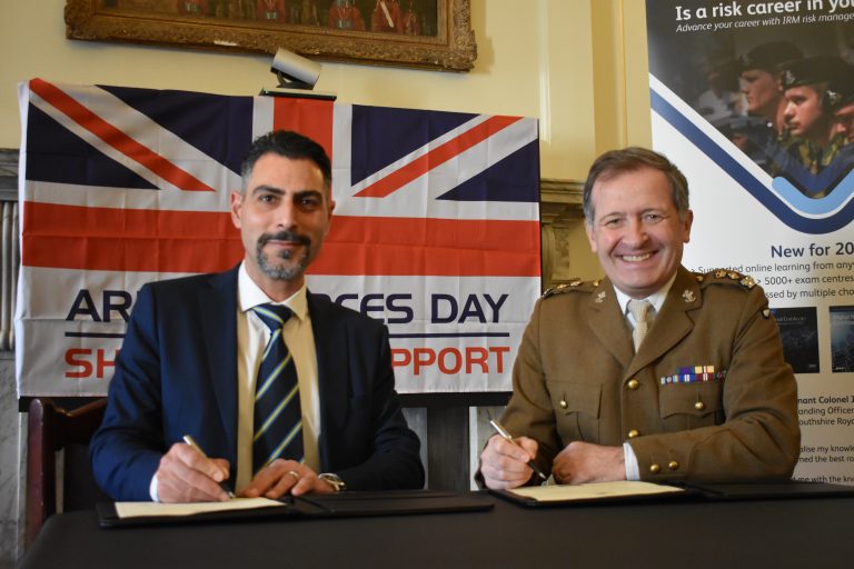 Two men seated at a table with pens, one in a suit and tie, the other in military uniform. A UK flag and Army Forces Day sign are in the background as they smile, celebrating the Armed Forces Covenant.