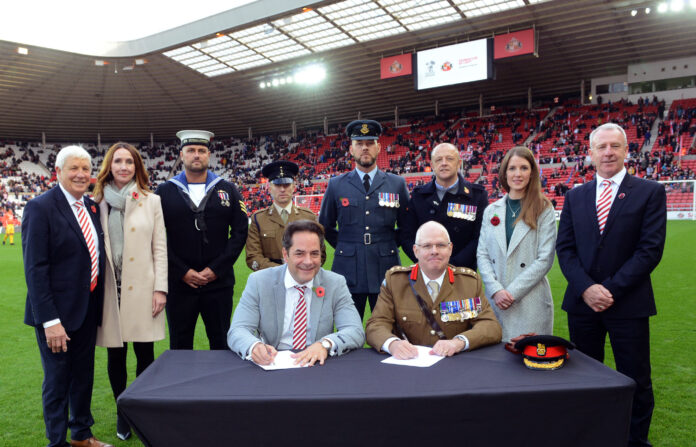 On a football field, under the vigilant eye of military personnel and civilians alike, two individuals sit at a table, signing documents to seal the Armed Forces Covenant in partnership with Foundation Of Light and Sunderland AFC.