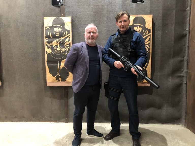 Two men stand indoors; one in a suit and the other, resembling Tommy Atkins, holds a firearm while clad in tactical gear. Two target posters hang in the background, reminiscent of Steve Heaney's operations in Sierra Leone.