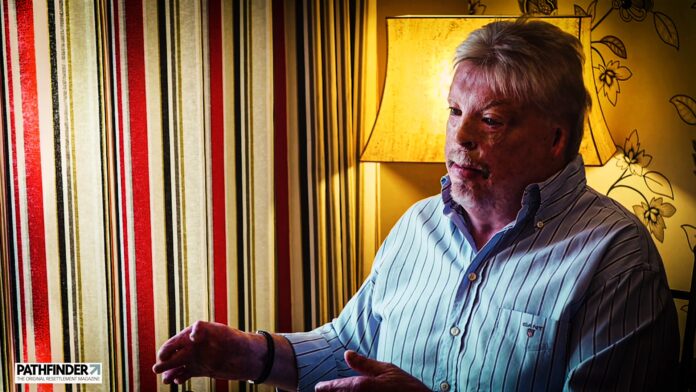 An older man with gray hair speaks, gesturing with his hands in a room adorned with striped curtains and a lit lamp. The dignity of a Falklands veteran resonates in his voice, echoing tales of courage and resilience.
